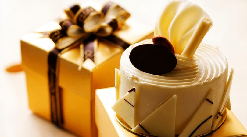 Elegant white cake with chocolate decorations next to a golden gift box with a bow.