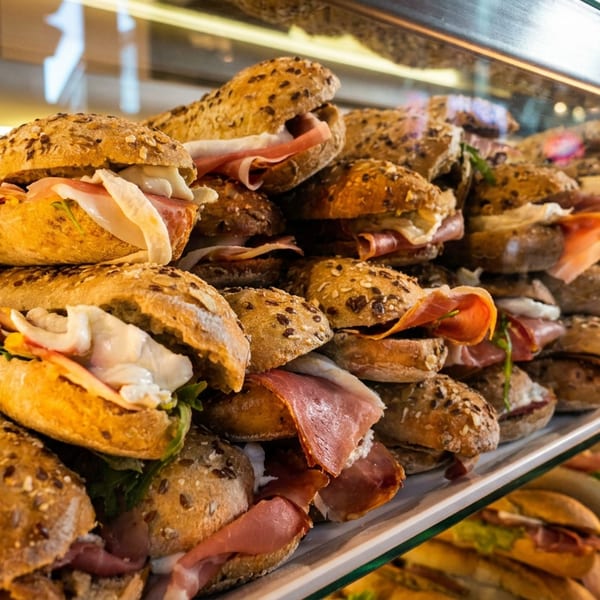 Abundant display of fresh sandwiches and rolls with prosciutto, cheese, and greens in a glass case.