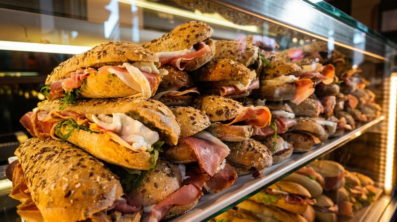 Rows of delicious sandwiches with ham, cheese, and arugula in a brightly lit display case.