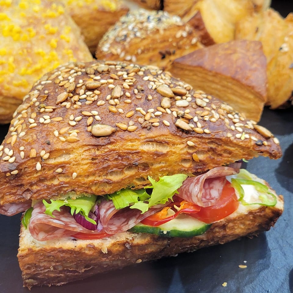 Seeded croissant sandwich filled with salami, lettuce, tomato, and cucumber on a dark surface with other pastries.