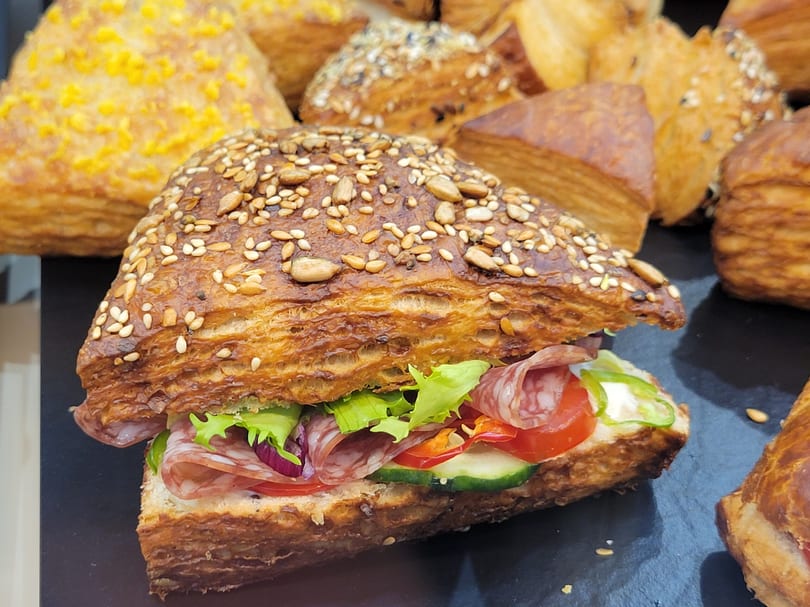 Seeded croissant sandwich filled with salami, lettuce, tomato, and cucumber on a dark surface with other pastries.