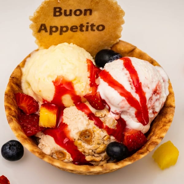 Three scoops of ice cream in a waffle bowl with berries, mango, and a "Buon Appetito" cookie.